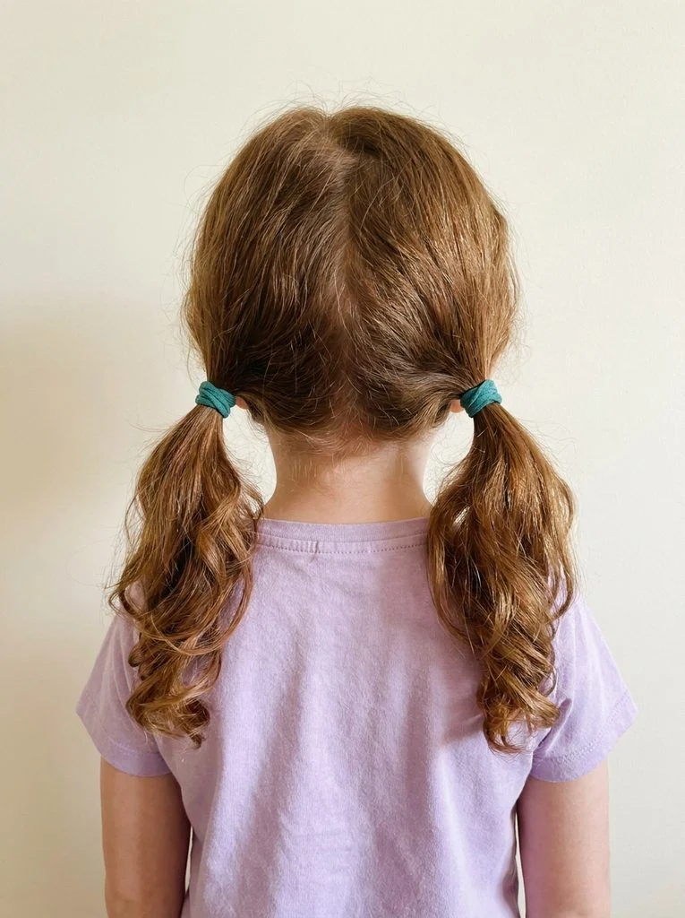 Two low pigtails on wavy auburn hair with matching teal ties.