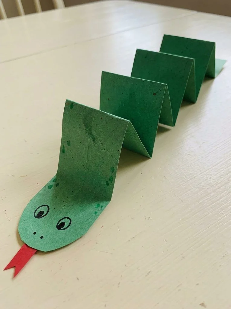 Green paper snake with an accordion body, drawn spots, and a red paper tongue.