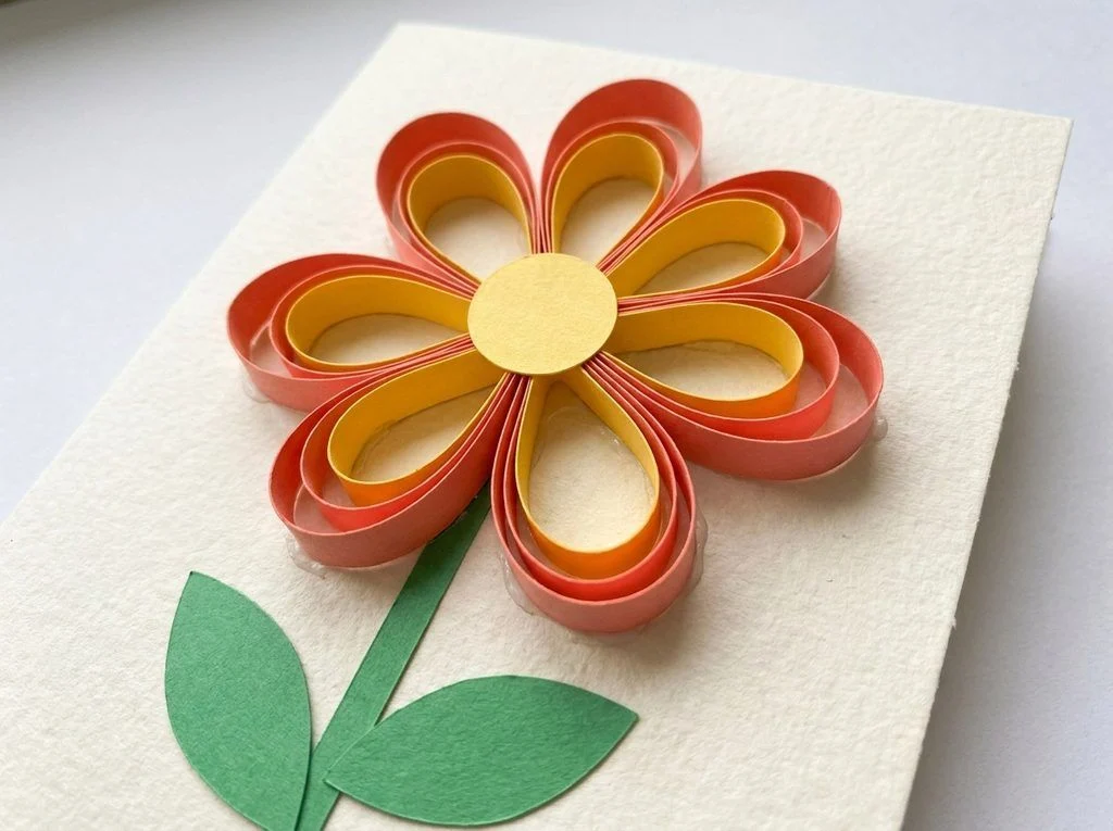 Paper flower with raised loop petals, a yellow center, and a flat green stem.