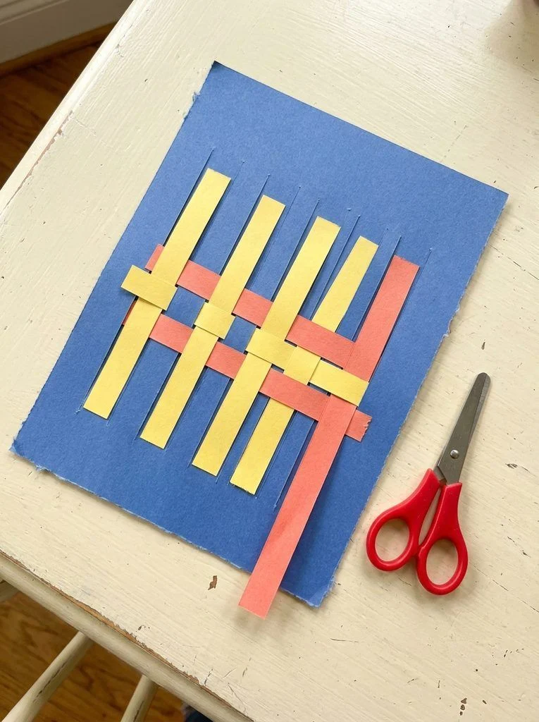 Colored paper strips woven through a blue slotted sheet in an over-under pattern.